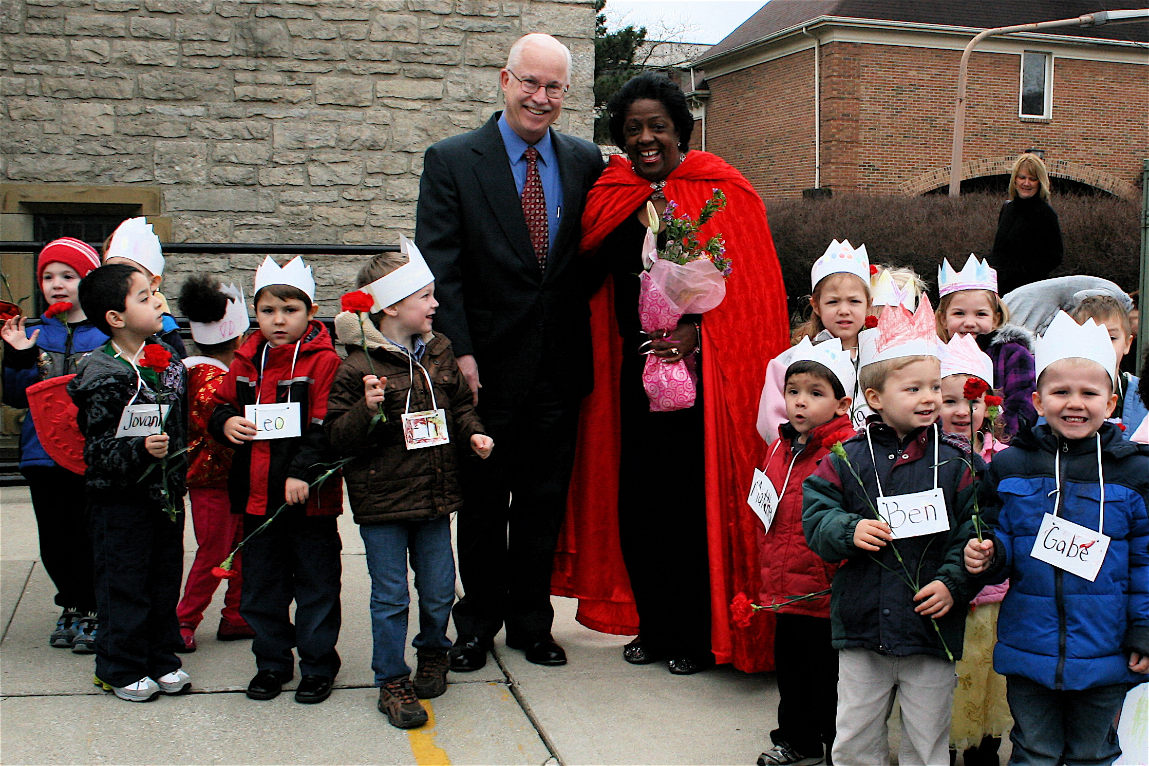 Jed Morison and Becky Love standing outside with many young children. Becky is wearing a red cape.