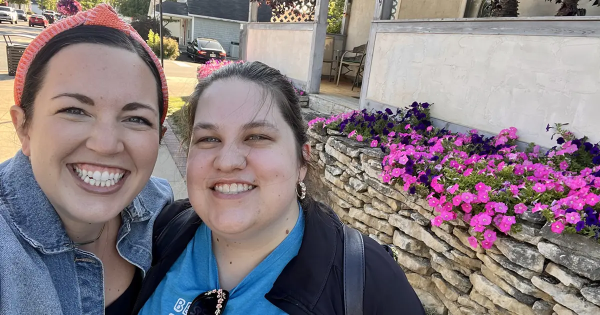 Two women smiling in near a flower bed with purple flowers