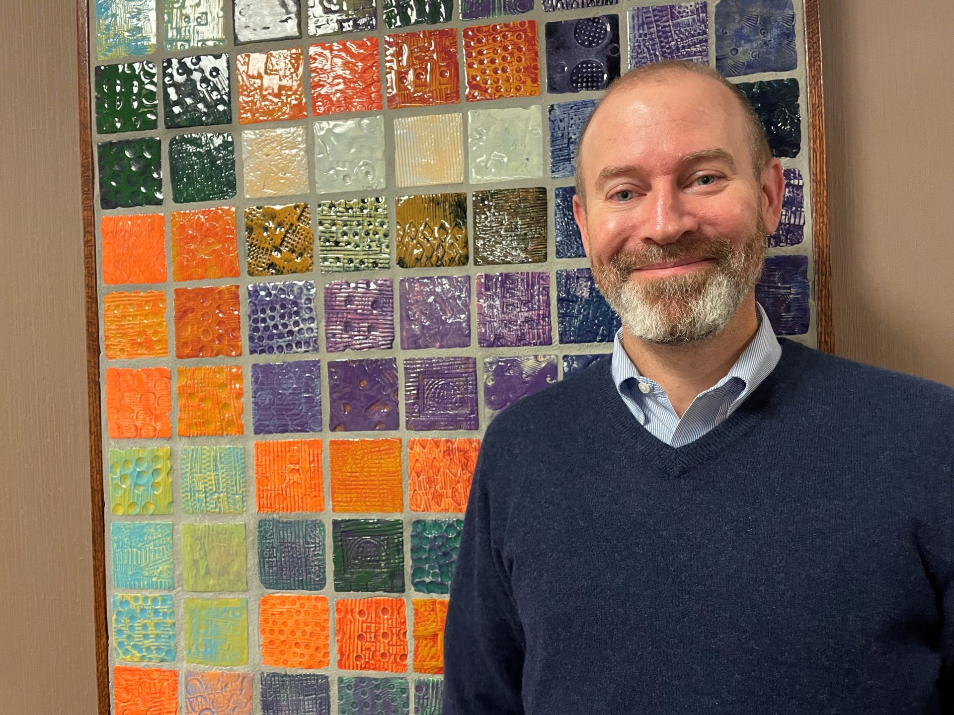 Smiling man with beard standing in front of colorful mosiac.